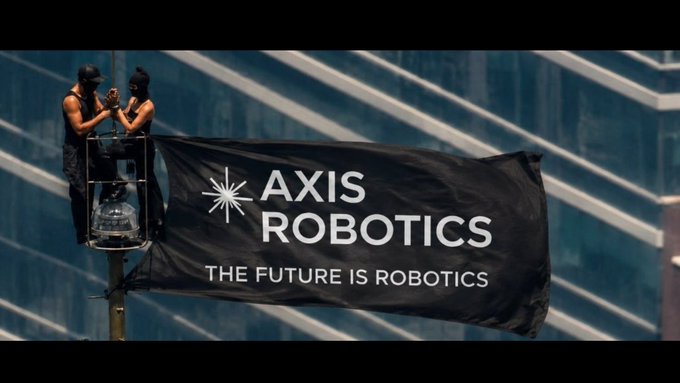 Two people in ski masks standing on a platform installing a large black banner that reads 'AXIS ROBOTICS - THE FUTURE IS ROBOTICS' on a modern glass building.