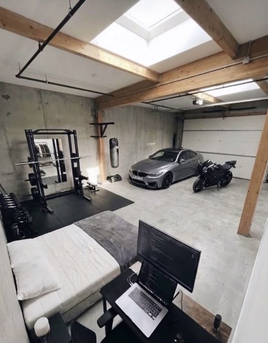 An interior view of a multi-purpose garage space containing a bed, a squat rack with weights, a desk with dual monitors, a silver BMW, and a motorcycle.