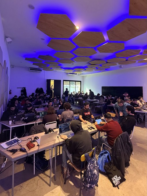 A photograph of a crowded hackathon or co-working space with people working on laptops at long tables, illuminated by striking blue lighting from hexagonal wooden panels on the ceiling.