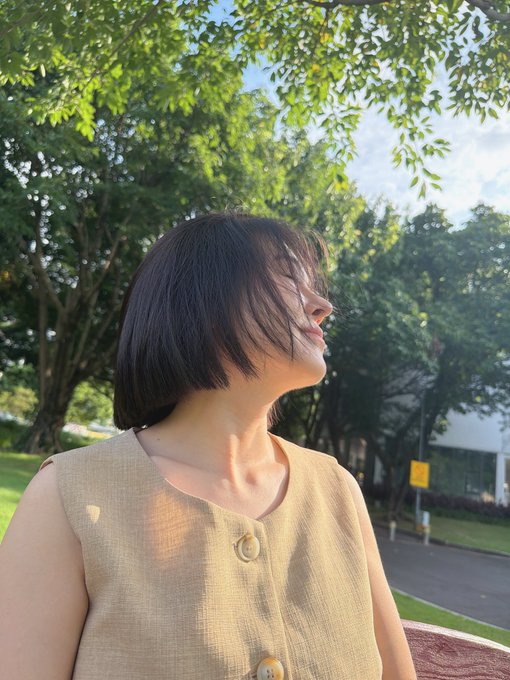 A woman with a sleek bob haircut is shown in profile, basking in the golden afternoon sunlight under leafy trees, wearing a beige sleeveless top. The warm, candid portrait captures a relaxed, joyful moment that aligns with the post's tone of mutual appreciation between friends—one who introduced her to Pi Network cryptocurrency and another who treats her to meals in exchange for professional photos.