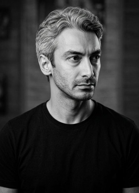 Black-and-white portrait of a man with light hair and stubble wearing a black t-shirt, looking off to the side.