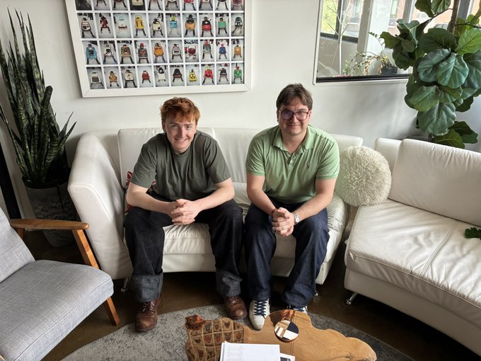 Two young men sit on a white couch in a living room with large plants; framed artwork showing various characters hangs on the wall behind them.