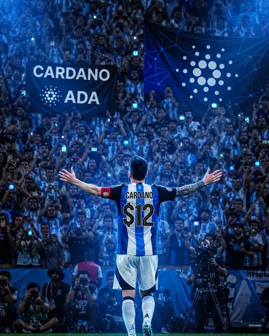 A soccer player stands with arms outstretched facing a packed stadium crowd, wearing a blue and black striped jersey with "CARDANO" and "$12" printed on the back, while large banners displaying "CARDANO ADA" and the Cardano logo are held up by fans in the stands, visually reinforcing the post's theme of Cardano's expanding "#ADA universe" through fan-driven promotion.