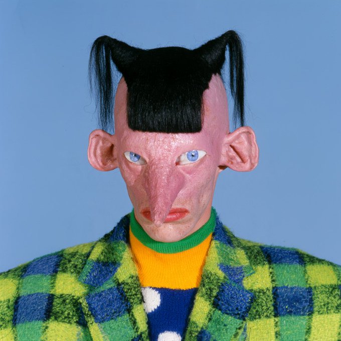 A grotesque puppet figure with pink skin, two black pigtails on a shaved head, a long pointed nose, and piercing blue eyes wears a vibrant green and yellow plaid jacket over a colorful patterned shirt, perfectly embodying the eerie-yet-playful "creeptopunksssss" aesthetic with its punk-inspired yet unsettling design.