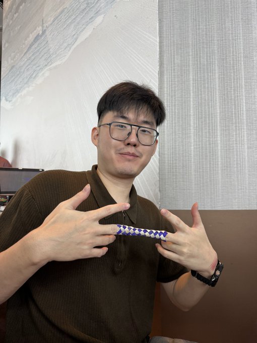 A young man wearing glasses and a brown polo shirt holding a blue and white checkered object, posing with hand gestures.