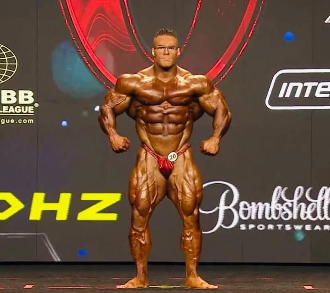 A heavily muscled male bodybuilder in red posing trunks standing on a competition stage with IFBB and Bombshell Sportswear logos in the background.