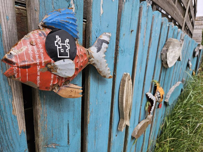 The image features a blue wooden fence with various fish decorations hanging from it, including one large fish-shaped decoration and several smaller ones. The fish decorations are made of metal or wood and are attached to the fence using nails or screws. The post in the image reads "GM @Eldorim_xyz loading". This suggests that the image is related to a social media platform where users can share photos, videos, and other content with their friends and followers.