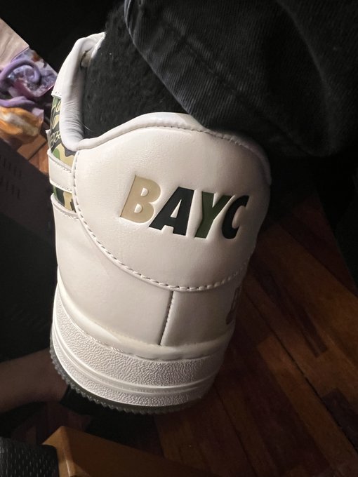 A close-up of a white sneaker featuring the gold 'BAYC' (Bored Ape Yacht Club) logo on the heel, worn with dark pants.
