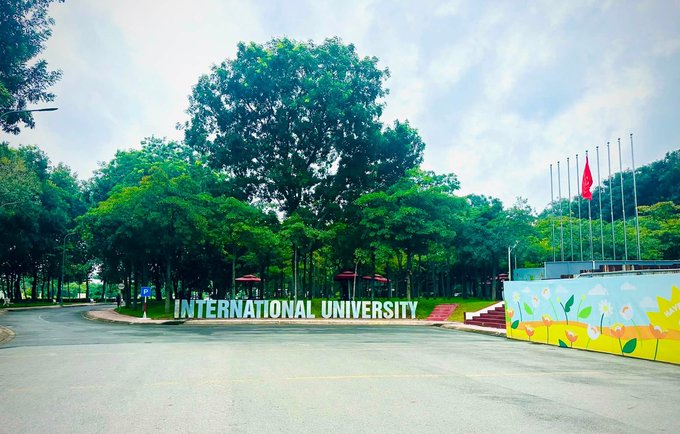 Photograph of an entrance to 'INTERNATIONAL UNIVERSITY' with a colorful mural, flagpoles flying a red flag, and lush green trees lining the road.