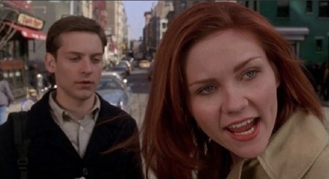 A cinematic still from the movie Spider-Man featuring Tobey Maguire and Kirsten Dunst on a busy city street, with Dunst in the foreground speaking and Maguire in the background.