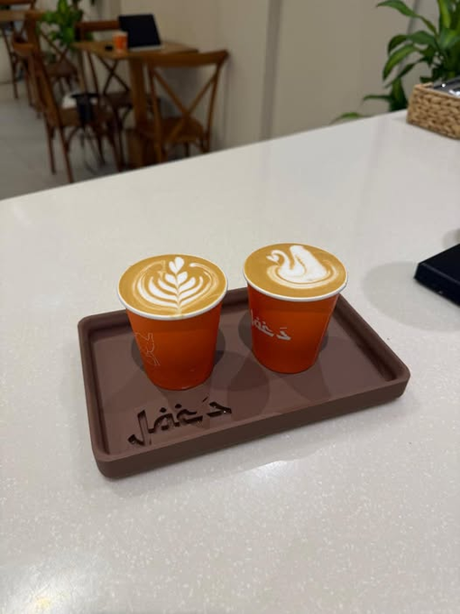 Two orange paper cups with latte art sit on a brown tray bearing Arabic script, placed on a white counter.