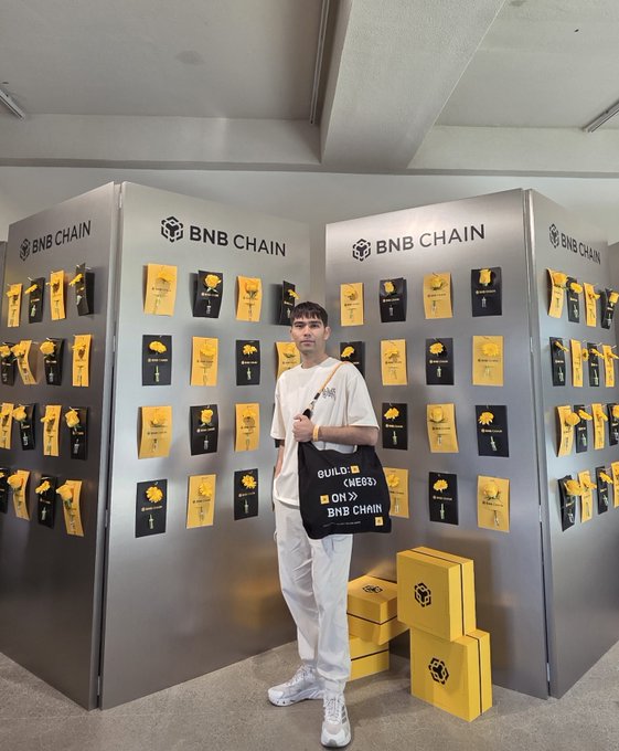 A young man wearing a white t-shirt and holding a black BNB Chain bag stands in front of a display featuring BNB Chain logos and yellow sunflower cards.