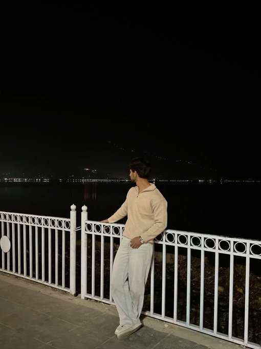 A man in a beige sweater and light pants leans against a white metal railing at night, gazing out over a dark body of water with distant city lights and mountains visible on the horizon.