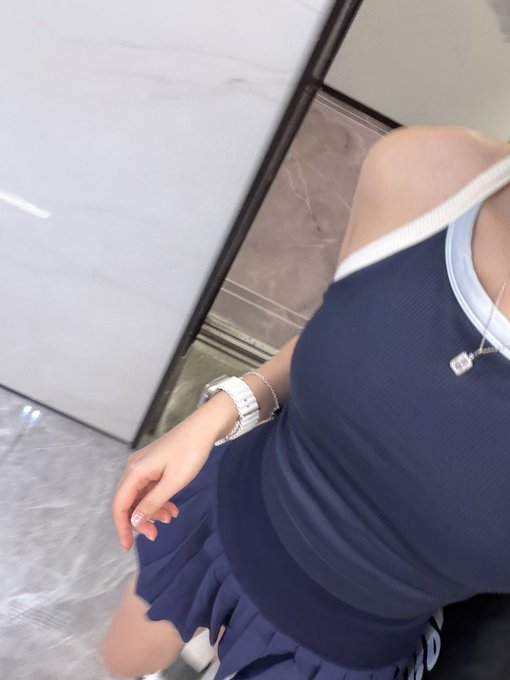 A selfie-style photo showing a person's torso in a navy blue dress with white trim, wearing a watch and necklace, against a marble-like wall.