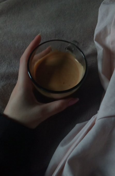 A hand holding a glass mug filled with a brown beverage, viewed from above against a dark gray surface next to a pink fabric.