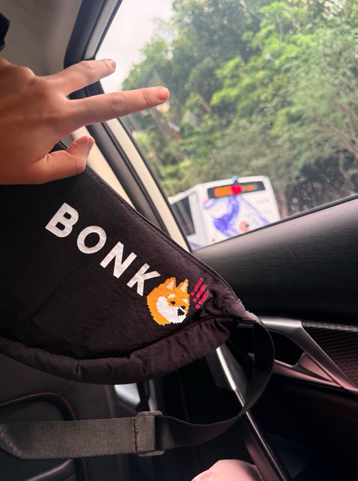 A hand makes a peace sign from inside a car, with a black "BONK" branded bag featuring a Shiba Inu dog logo visible in the foreground and trees seen through the window.