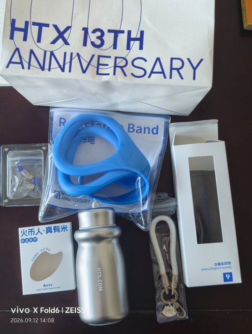 HTX 13th Anniversary merchandise items spread out including a paper bag, blue resistance bands, a stainless steel HTX.COM water bottle, an 'Au' (Gold) tag, keychains, and other branded items, photo taken with a vivo X Fold6.