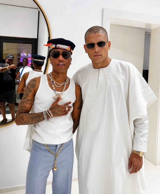 Two men pose together indoors; the one on the left wears a white tank top, blue pants, layered pearl necklaces, sunglasses, and a cap, while the taller man on the right wears a flowing white robe and sunglasses.