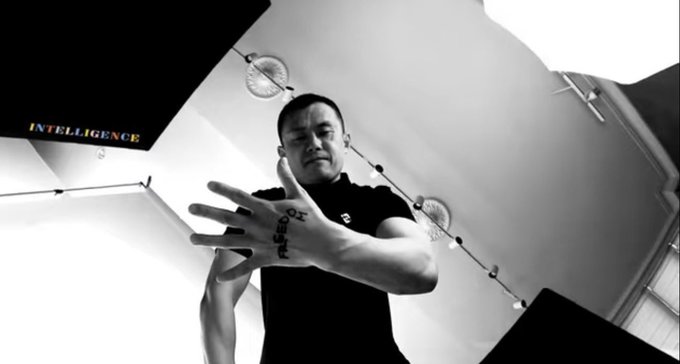 A black and white photo of a man with a hand tattoo extending his fingers toward the camera, with 'INTELLIGENCE' visible on the wall behind him.