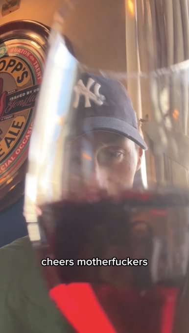 A first-person selfie through a wine glass with red wine, showing a person wearing a New York Yankees cap behind the glass, with a bottle of Newcastle Brown Ale in the background; text overlay reads 'cheers motherfuckers'.