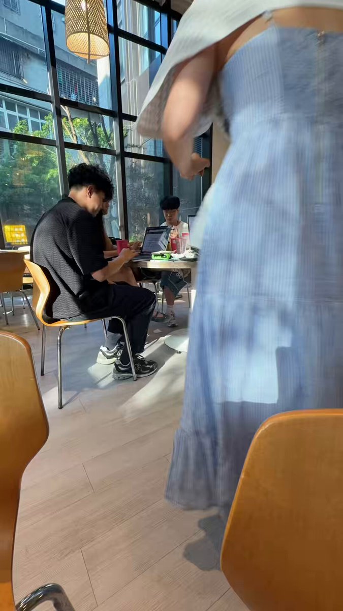 The image shows a man sitting at a table with his laptop, while another person sits across from him. A third person can be seen standing behind them. The scene takes place in an office or cafe setting, where people are working on their laptops.