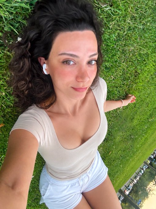 A woman with curly brown hair wearing a beige scoop-neck top and white shorts takes an overhead selfie while lying on lush green grass with white wireless earbuds in. The caption "Gm frens! It's Monday. Touch some grass today" pairs perfectly with the image as she literally "touched grass" by relaxing outdoors on a sunny day.
