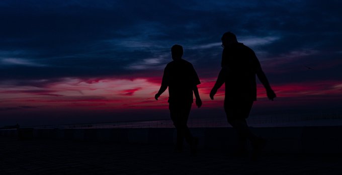 Silhouettes of two people walking against a dramatic sunset sky with deep red and dark blue clouds.