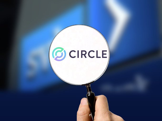 A close-up of a hand holding a magnifying glass focused on the Circle (cryptocurrency company) logo, with a blurred blue screen in the background.