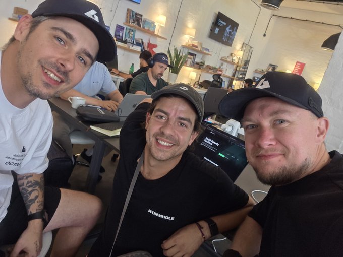 A selfie of three smiling men in a casual co-working space, wearing caps and dark clothing, with laptops and office furniture visible in the background.