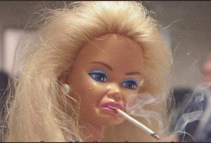 A close-up of a blonde doll-like figure with blue eyeshadow, pink lips, and a cigarette in its mouth, with smoke visible.