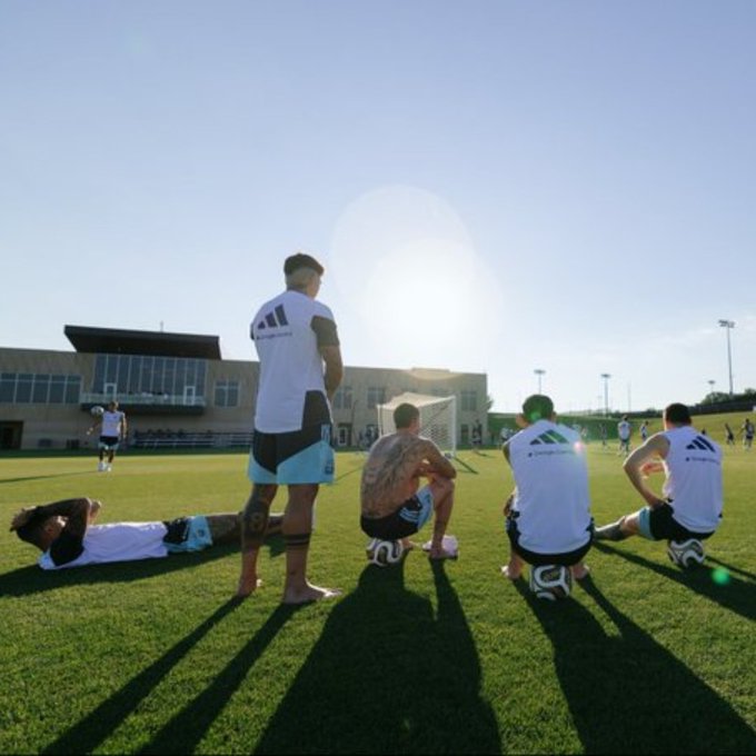 Soccer players in white training jerseys gather on a sunlit practice field near a goal, with one player stretching on the grass while others kneel or stand, casting long shadows in the late afternoon light. The image reflects a relaxed team training moment, supporting the post's call to "drop to show support" for the squad and community.