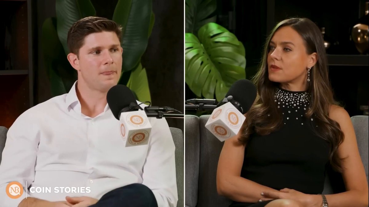 In this image, there are two people sitting next to each other with microphones in front of them. They appear to be having a conversation or participating in an interview. The background features a plant, which adds some greenery to the scene. The main focus is on the two individuals and their interaction with the camera equipment.