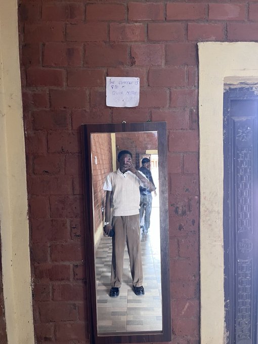 A man's reflection appears in a tall mirror mounted on a red brick wall, showing him taking the photo in a hallway with another person behind him; a handwritten note is posted above the mirror.