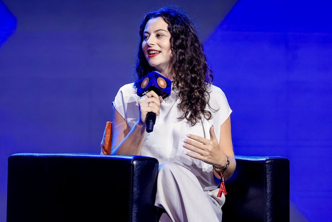 A woman with long curly dark hair, wearing a white blouse and red lipstick, speaks into a blue microphone bearing orange Bitcoin "₿" logos while seated on a stage with a blue backdrop. The post "The world will know about Bitcoin and I'll be one of the voices to tell them" aligns with her role as a Bitcoin advocate amplifying awareness of the cryptocurrency through public speaking.