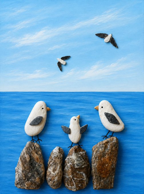 A painted pebble craft scene of three seagulls made from white and grey stones perched on rock formations against a blue sky and ocean backdrop, with two flying birds in the distance.