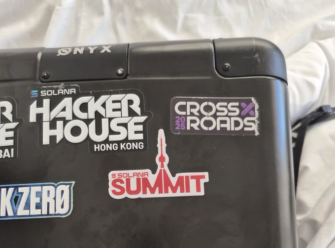 A black laptop covered in stickers, including 'ONYX', 'SOLANA HACKER HOUSE HONG KONG', 'CROSS ROADS 2025', 'SOLANA SUMMIT', and a 'STARK ZERO' sticker.