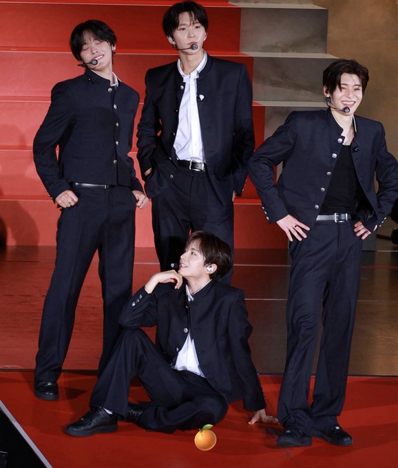 Four young men in matching black military-style uniforms with white buttons pose on stage, three standing and one seated on the red platform, with what appears to be a digitally added orange emoji near his foot—likely referencing the "airdropped" item mentioned in the caption.