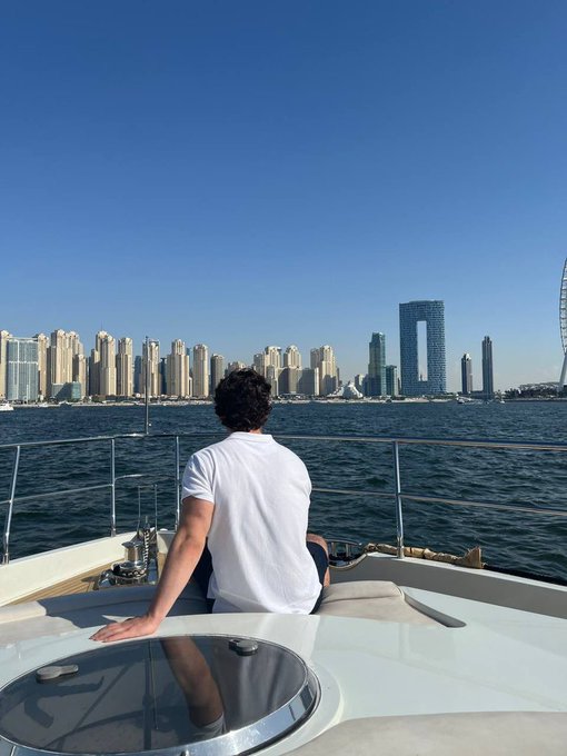 A young man in a white t-shirt sits on the bow of a yacht looking out at the Dubai skyline, with the Ain Dubai Ferris wheel visible on the right.