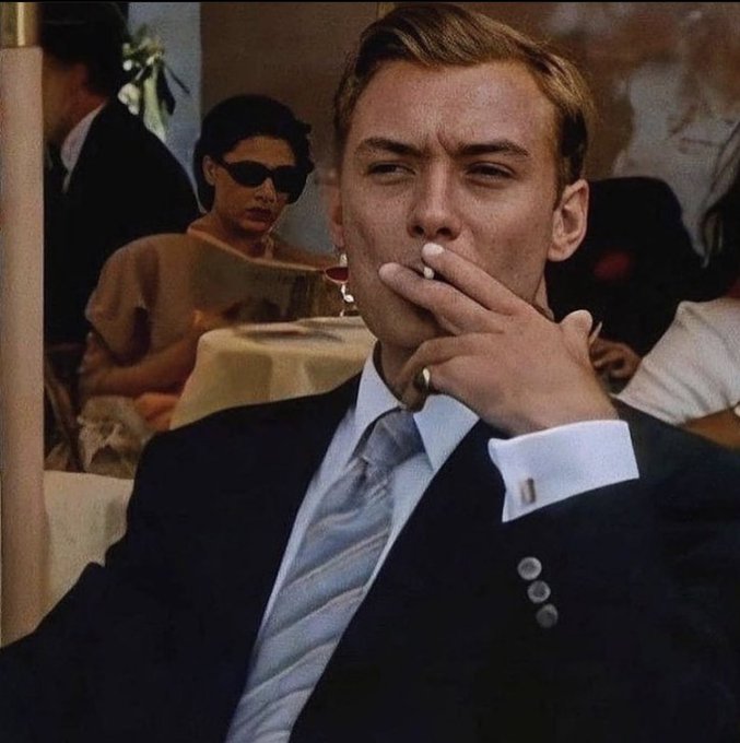 A cinematic scene of a blonde man in a dark suit smoking a cigarette, with a woman wearing sunglasses visible in the background at what appears to be a restaurant.