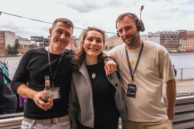 Three people pose together at what appears to be the Solana Summit during Berlin Blockchain Week, with the man on the right wearing a "SUPERTEAM" lanyard and headset while holding the woman in the middle, and the man on the left holding a beer bottle. The image ties to the post as a candid moment captured by @Son_of_a_Pigeon before the first day of BBW concluded, celebrating SuperteamDE's role in kicking off the event.