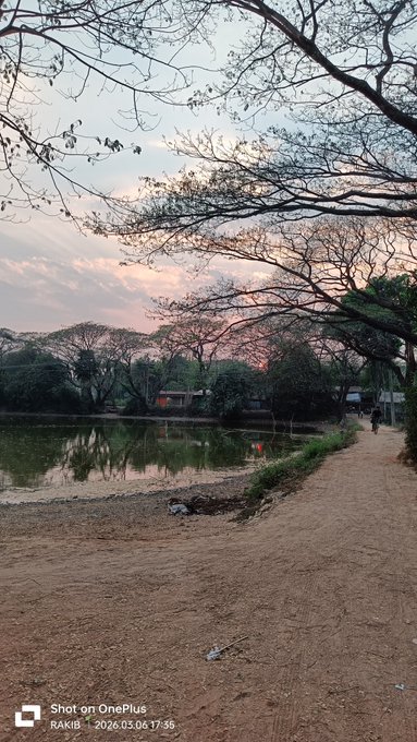A landscape photograph taken at dusk showing a dirt path next to a calm pond reflecting bare trees and a pinkish sky, with a camera watermark reading 'Shot on OnePlus RAKIB | 2026.03.04 17:36' in the bottom left corner.