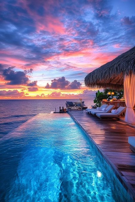 The image features a beautiful beachfront property with an outdoor swimming pool that overlooks the ocean at sunset. The sky above the water has a stunning pink hue, creating a picturesque scene. There are several chairs placed around the pool area for relaxation, and a dining table nearby. A person can be seen sitting on one of these chairs, enjoying the view or taking in the ambiance.