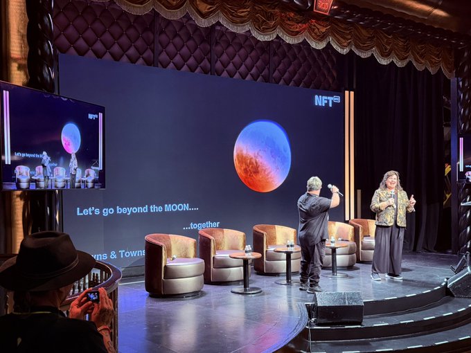 Two speakers stand on a stage in front of a large screen displaying a red planet and the text "Let's go beyond the MOON...." and "NFT" during a conference panel.
