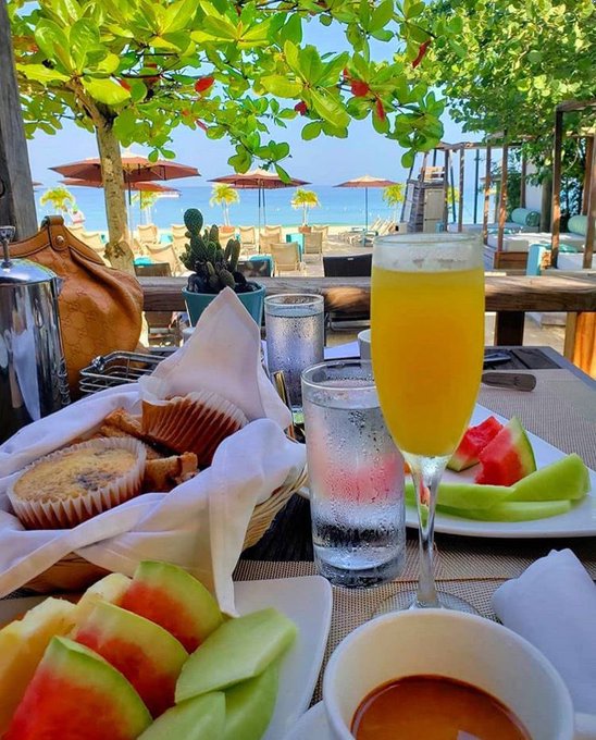 The image shows a table with various food items such as fruit, muffins, and water glasses. There are also two wine glasses filled with orange juice placed on the table. The scene appears to be set at an outdoor restaurant or cafe overlooking the ocean. The focus of the image is on the food and drinks served, creating a relaxed atmosphere for diners enjoying their meal by the beach.