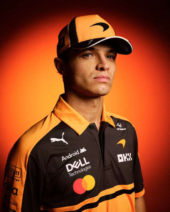 A young man models a McLaren F1 team polo shirt featuring sponsor logos including Android, Dell Technologies, OKX, Puma, and Hilton, paired with an orange McLaren cap. The bold orange and black team colors align perfectly with a summer motorsport aesthetic, making this a stylish warm-weather look for racing fans.