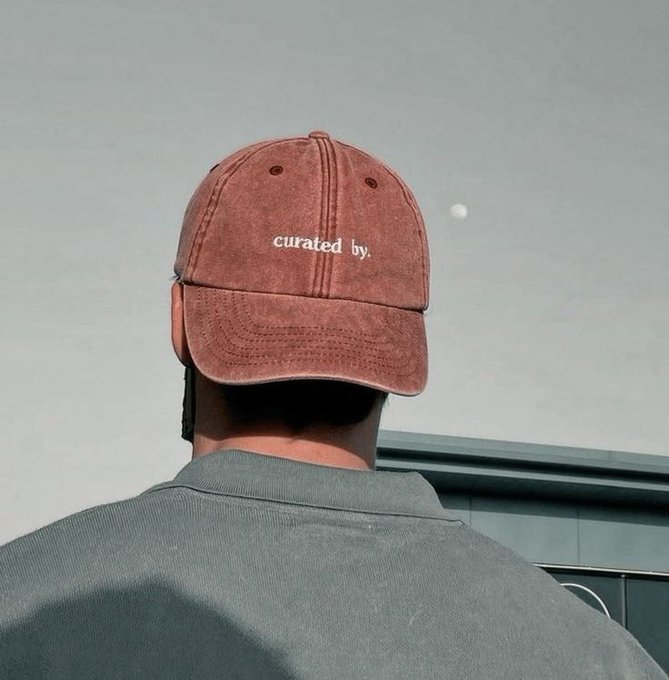 A person viewed from behind wears a faded red baseball cap with the text "curated by." embroidered in white, paired with a teal polo shirt against a muted sky background.

The cap's "curated by" text metaphorically aligns with the post's message about selecting better, more curated blockchain infrastructure solutions rather than settling for congested standard networks that bottleneck during high-demand events.