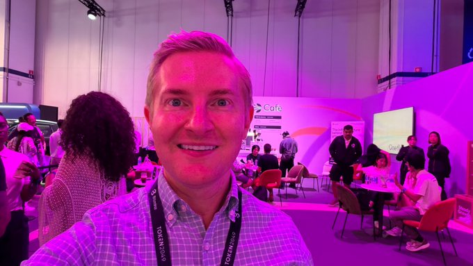 A smiling man with a Token2049 lanyard takes a selfie at a conference event with pink lighting and attendees visible in the background.