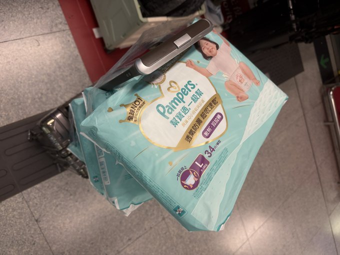 A package of **Pampers 幫寶適 一級幫** diapers (size L, 34 pieces) rests on a suitcase handle, with the post's caption comparing the dad's quick dash to Hong Kong for diapers to on-chain engineers swiftly supporting hot projects—suggesting he's travel-ready for both parenting and crypto.