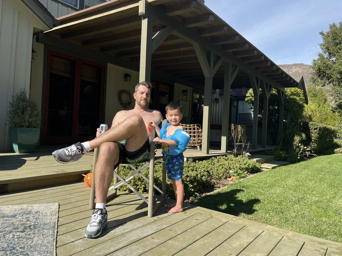 A casual outdoor photo showing a shirtless man sitting in a folding chair on a wooden deck, with a young boy standing next to him wearing blue inflatable armbands and swim trunks.