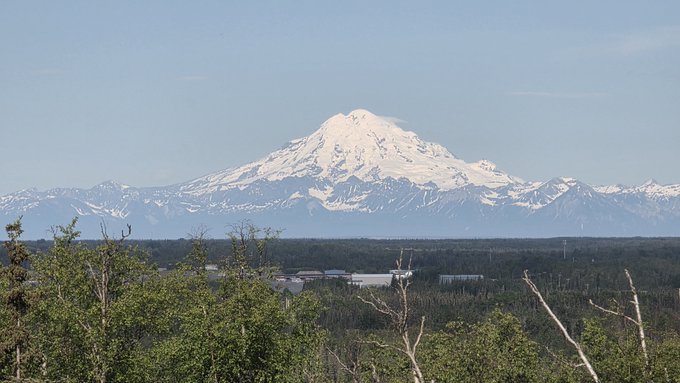 A snow-capped mountain peak rises prominently above a forested landscape under a clear blue sky.